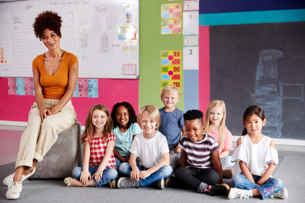 Portrait Elementary School Pupils Sitting On Floor In Classroom With Female Teacher